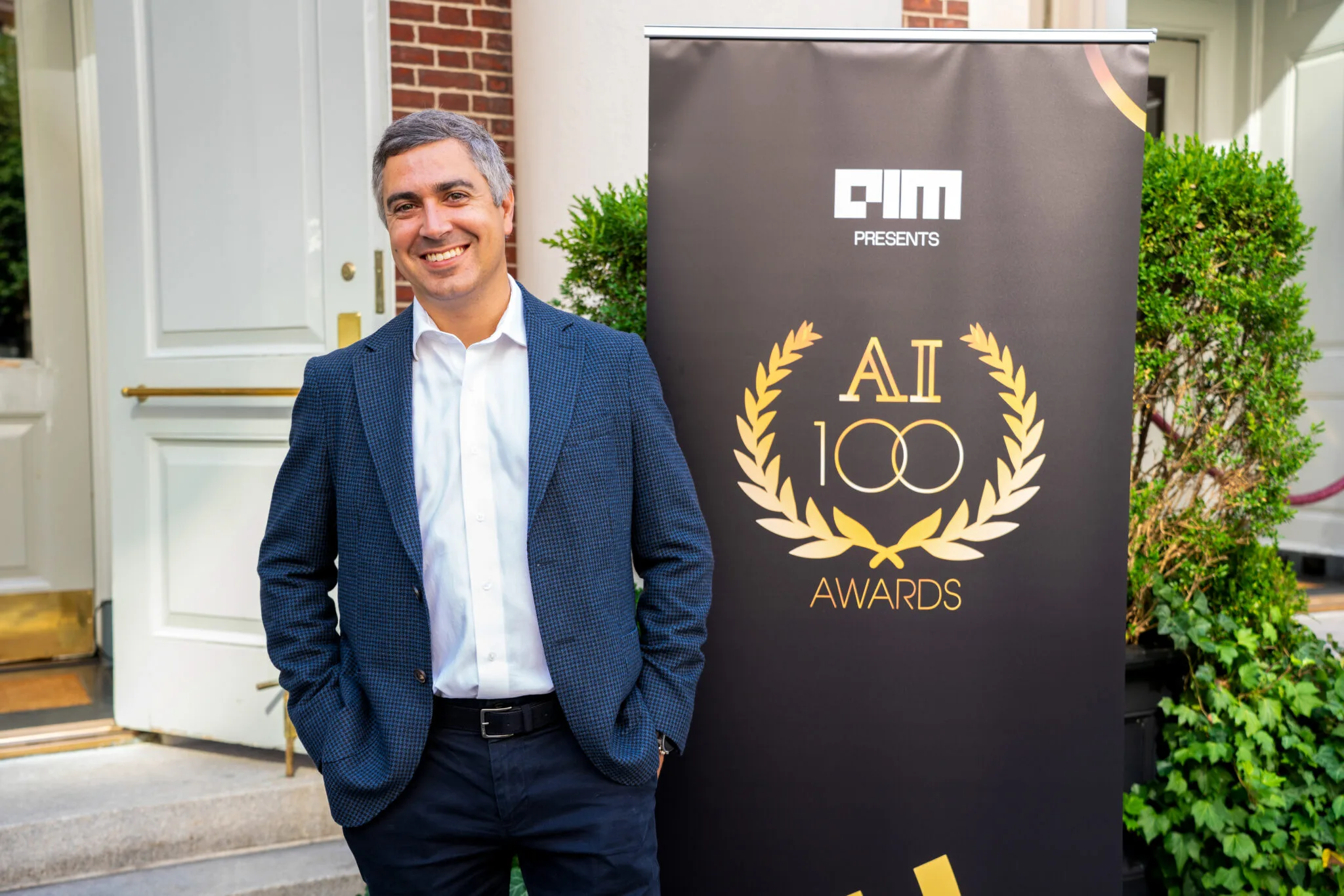 Smiling man by AI100 banner at MachineCon New York 2025